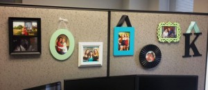 A beige office cubicle wall decorated with six framed photos of people, mostly women, and a large decorative letter K hanging on the right side.