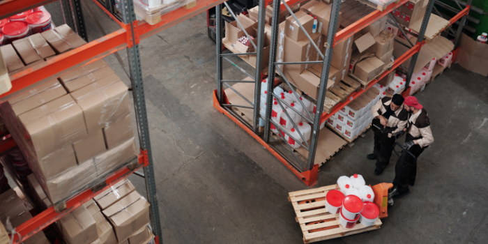 Aerial view of two workers in uniforms and masks standing next to a pallet with red and white buckets in a warehouse filled with shelves of boxes and containers.