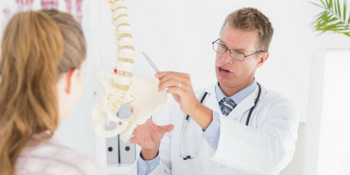 A doctor in a white coat and glasses, similar to many chiropractors, explains a human spine and pelvis model to a seated patient in a medical office.