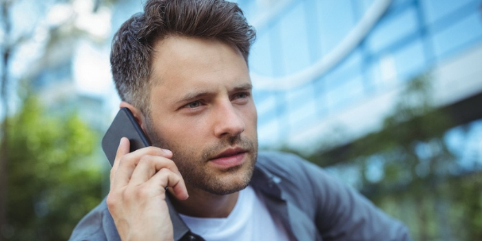 A young man with short brown hair is holding a smartphone to his ear, appearing to have a conversation outdoors in front of a modern glass building.