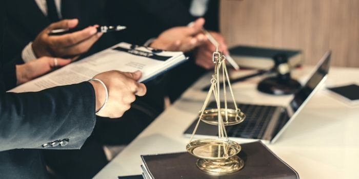 Two people in business attire discuss documents at a desk with a laptop. A golden justice scale sits on top of a book in the foreground, symbolizing law and legal matters.