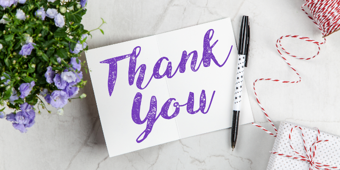 A card with the words Thank You in purple script sits open on a marble surface beside a black pen, a small potted plant with purple flowers, and a white gift box wrapped with red and white twine.