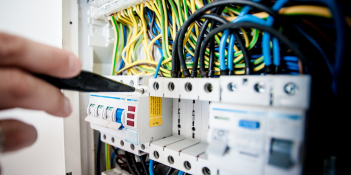 A hand uses a tool to adjust or inspect a circuit breaker in an electrical panel filled with colored wires and switches.