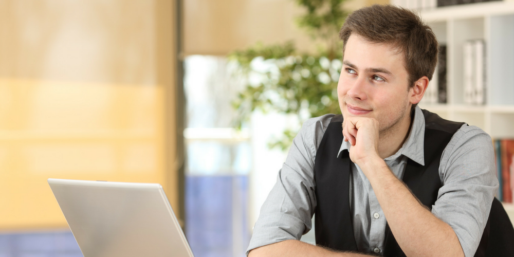 A young man sitting at a desk with a laptop, resting his chin on his hand and looking thoughtfully into the distance, with shelves and a plant in the background.