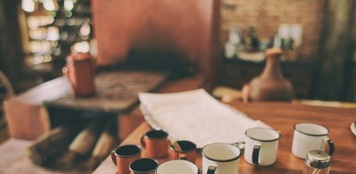 A wooden table with several enamel mugs and cups in white and brown, along with a tray and cloth, sits in front of a rustic kitchen—perfect for gatherings supporting the Animal Foundation, with shelves and a brick wall in the background.