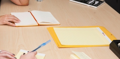 Two people sitting at a light wooden table with notebooks, a yellow legal pad, a pen, and a closed envelope, engaged in a meeting or discussion.