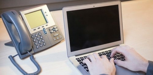 A person types on a laptop keyboard at a desk with a corded office phone beside the computer.