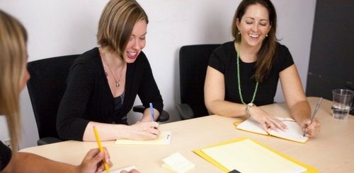 Three women sit at a conference table, smiling and writing in notebooks during a meeting. There are folders, papers, and a glass of water on the table.