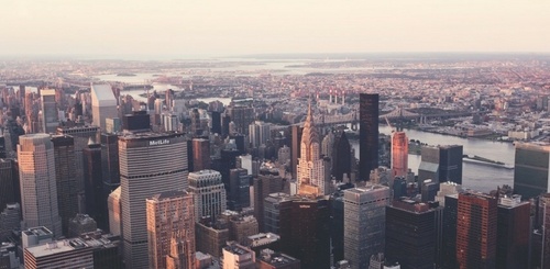 Aerial view of a city skyline with many tall skyscrapers, a river running through the background, and a hazy sky at sunset.