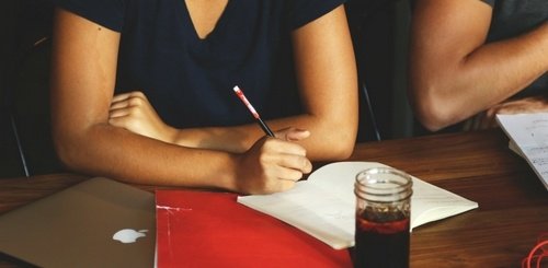 Two people sitting at a wooden table with a laptop, red folder, open notebook, and a jar of dark beverage. One person is writing in the notebook with a pencil. Only their arms and torsos are visible.