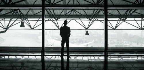 A person stands alone, silhouetted against large windows at an airport terminal, perhaps awaiting a flight after using a Business Answering Service, gazing out at a plane on the tarmac on a foggy day.