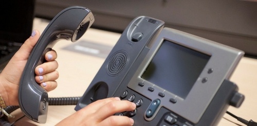 A person with light purple nail polish holds a handset and dials numbers on a gray office desk phone with a screen, buttons, and a coiled cord.