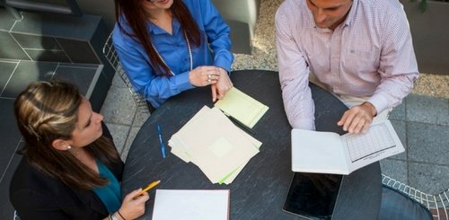 Three people sit around a round table with papers, a notebook, pens, and a digital tablet, engaged in discussion or collaboration, viewed from above.