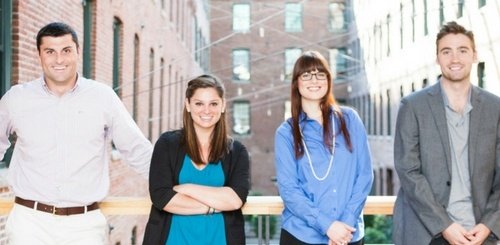 Four young adults, two men and two women, stand casually outdoors in a sunlit urban setting with brick buildings behind them, smiling at the camera—much like the teamwork seen when exploring the 3 crucial benefits of a virtual receptionist for law firms.