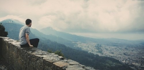 A person sits on a stone wall overlooking a city nestled among mountains, with a cloudy sky above and mist rolling over the distant hills.