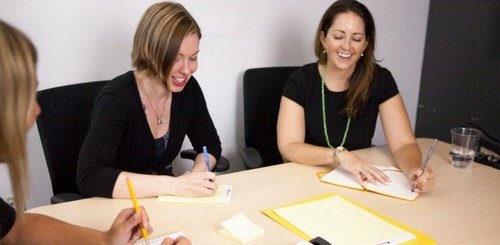 Three women sit at a table, writing in notebooks and smiling during a meeting. Notepads, sticky notes, and a glass of water are on the table. The atmosphere appears friendly and collaborative.