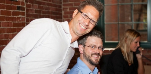 Two men smiling at the camera, one standing and one sitting, in a room with brick walls. A woman in the background is talking on the phone near a window.