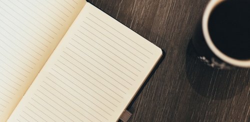 An open, lined notebook lies on a dark wooden surface next to a cup of black coffee. The scene suggests a setting for writing or note-taking.