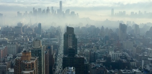 A wide view of a city skyline covered in light fog, with tall buildings fading into the distance and a hazy, overcast sky above.