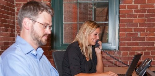 A woman talks on the phone while working on a laptop at a desk, smiling. A man with glasses sits nearby, focused on his own work. Both are in an office with exposed brick walls and a large window.