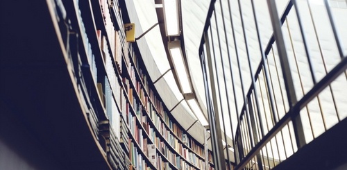Curved rows of bookshelves filled with books in a library, with a large window and a metal railing visible on the right side, allowing natural light to enter the space.