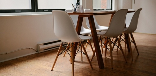A modern dining area with a wooden table and four white plastic chairs with wooden legs, set on a wooden floor near a large window letting in natural light.