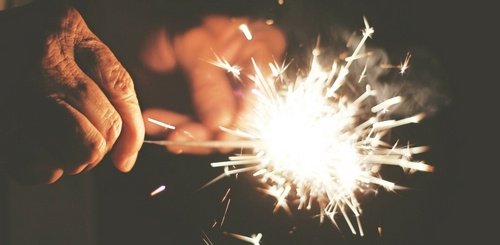 A close-up of a person’s hand holding a lit sparkler, with bright sparks and light illuminating the scene against a dark background.