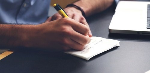 A person writes on a piece of paper with a yellow pen next to an open laptop on a dark desk.