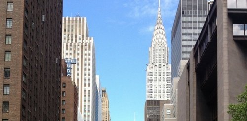 View of tall buildings in New York City with the Chrysler Building’s distinctive spire in the center background, under a blue sky with a few clouds.