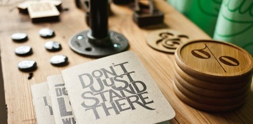 A wooden table with coasters, buttons, stamps, and a notebook in the foreground that reads DONT JUST STAND THERE in bold black letters.