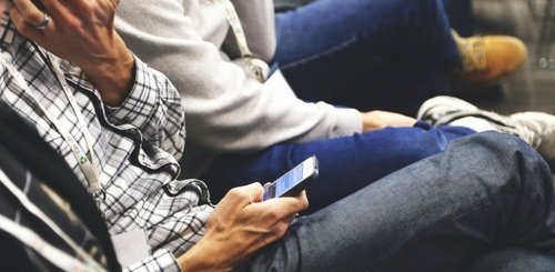 Two people sitting closely together, one wearing a plaid shirt, holding and looking at a smartphone, while the other person in a light-colored hoodie sits beside them with hands clasped on their lap.