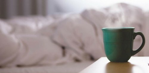 A green ceramic mug sits on a bedside table in the foreground, with an unmade bed and soft rumpled white bedding blurred in the background.