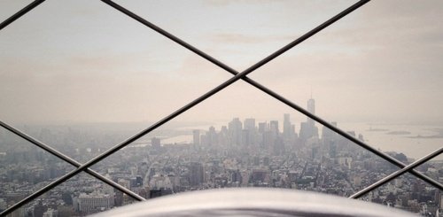 A hazy city skyline viewed through a metal safety grid, likely taken from a high vantage point such as an observation deck. Tall buildings and a distant body of water are visible under a cloudy sky.