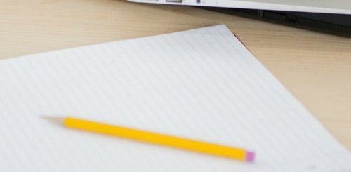 A yellow pencil rests on a lined sheet of paper with the corner of a laptop visible in the background on a light wooden desk.