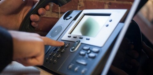 A person holds a phone receiver and presses buttons on an office desk phone with a digital display, set against a brick wall background, representing the professional touch provided by Abby Connect.