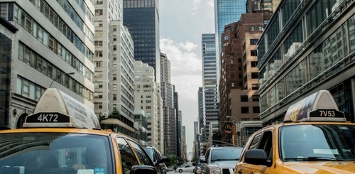 Yellow taxis drive down a busy city street lined with tall skyscrapers and office buildings. The sky is partly cloudy, and other vehicles are visible between the taxis.