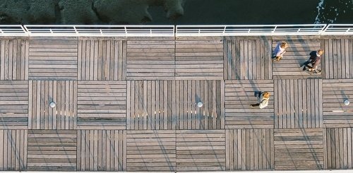 Aerial view of a wooden boardwalk with geometric plank patterns, bordered by a white railing. Three people walk along the path, casting long shadows in the sunlight. Water is visible at the top of the image.