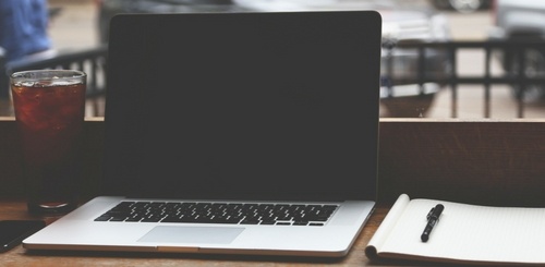 A closed laptop sits on a wooden table next to a glass of iced drink and an open notebook with a pen, suggesting a workspace in a café or casual setting.
