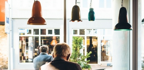 An older man sits alone at a table by a large window in a café, with pendant lights hanging above and a view of a street and storefronts outside during daylight.