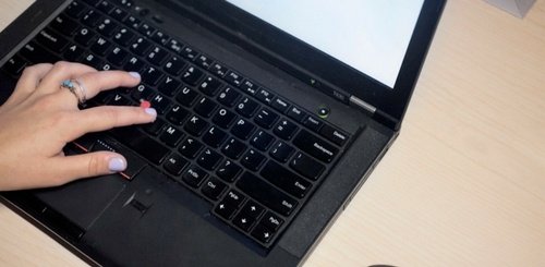 A person with light purple nail polish types on a black laptop keyboard with a blank white screen, on a light-colored wooden desk.