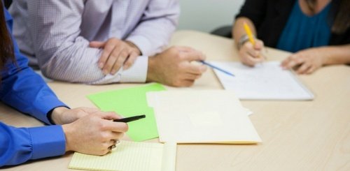 Three people sit at a table, discussing documents. Only their hands and arms are visible. They hold pens, with papers, folders, and notebooks spread out on the tabletop, suggesting a collaborative meeting.