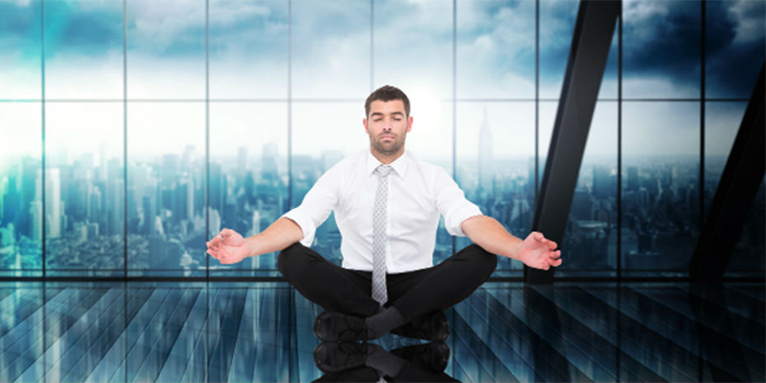 A man in business attire sits cross-legged, meditating with closed eyes in a modern glass office with a city skyline visible through the windows.