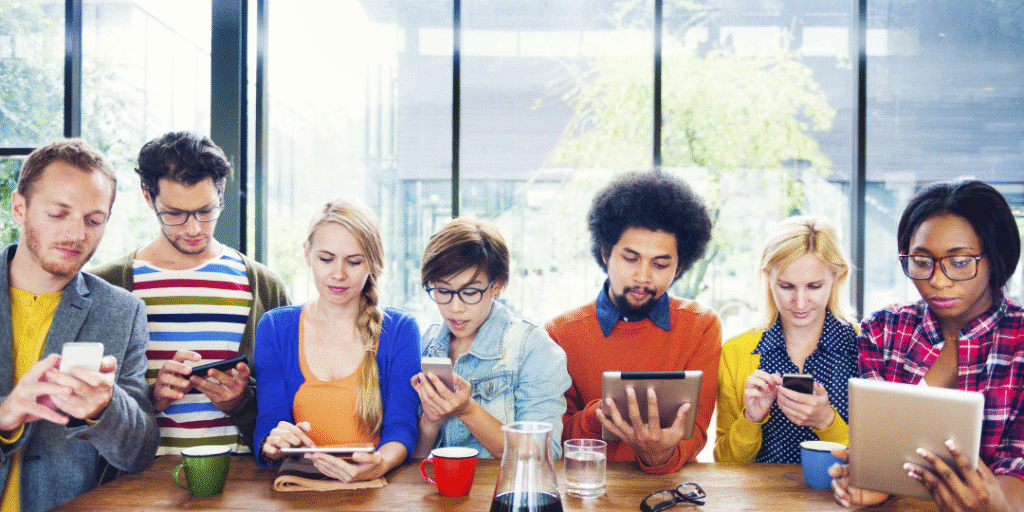 A diverse group of seven people sits at a long table by large windows, each focused on their smartphones or tablets. Coffee mugs and a glass pitcher are on the wooden table.