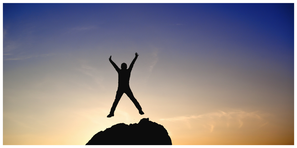 A person stands on a rock with arms and legs outstretched, silhouetted against a colorful sunset sky.