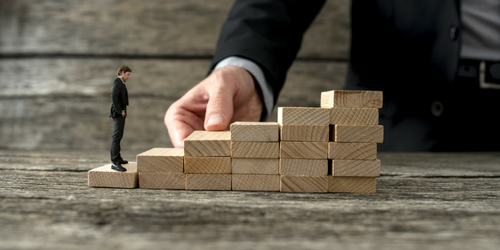 A large hand arranges wooden blocks as stairs while a tiny suited man stands at the bottom, contemplating the climb—symbolizing the journey to celebrate entrepreneurship. The rustic wood background adds depth to this inspiring scene.