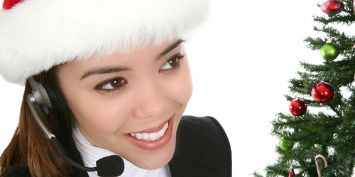 A woman wearing a Santa hat and a headset smiles while standing next to a decorated Christmas tree with red and green ornaments.