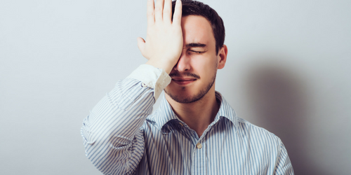 A man in a striped shirt stands against a plain background with his eyes closed and his palm pressed to his forehead, expressing frustration or realization.