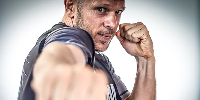 A man in a sports shirt poses in a boxing stance, with one fist extended toward the camera and the other near his face, looking intently forward against a plain, light background.