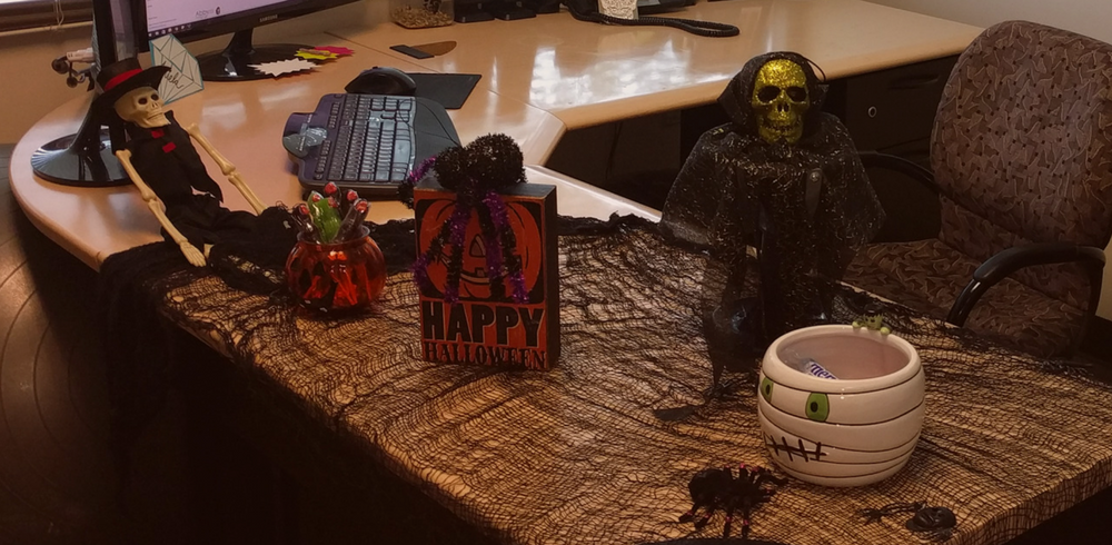 A desk decorated for Halloween with a skeleton doll, a golden skull with black cloth, a Happy Halloween sign, a pumpkin jar, a mummy bowl, plastic spiders, and black netting spread across the surface.