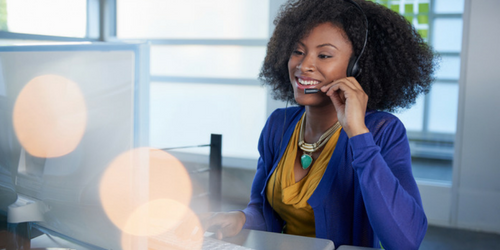 A woman wearing a headset smiles while working at a computer in an office setting. She is dressed in a yellow top with a blue cardigan and a statement necklace.